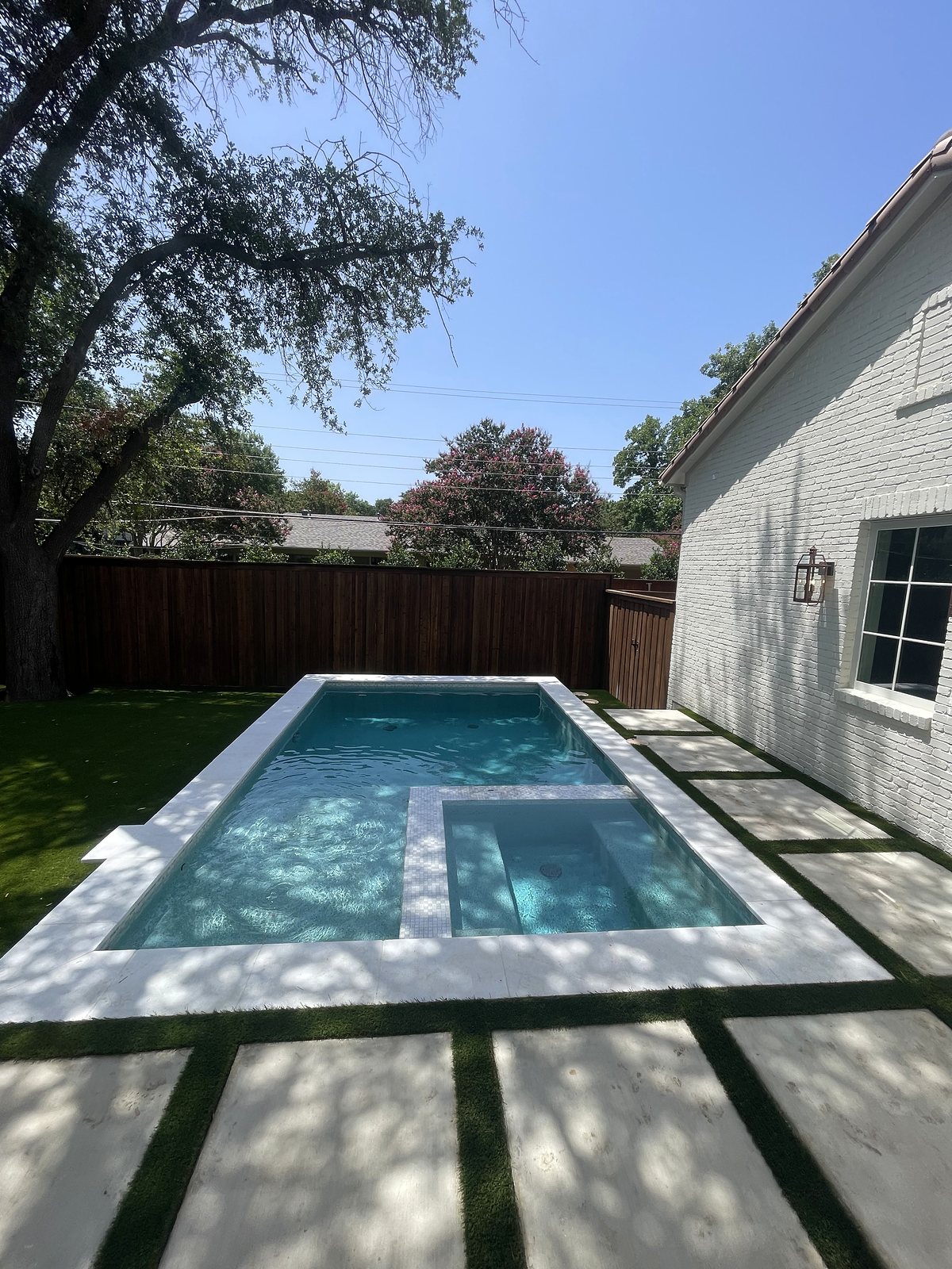 Pool and spa beside the white-brick home, framed by white coping and large pavers set in turf โ completed by Infinity Group DFW in Dallas, Texas